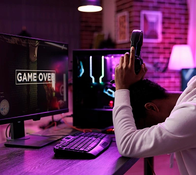 Een gefrustreerde gamer zit met zijn handen omhoog achter zijn computer nadat er tijdens het spelen een 'game over'-melding op het scherm verschijnt.