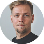 Professioneel portret van een jonge man met kort blond haar en een lichte baard, gekleed in een donker shirt, tegen een neutrale achtergrond.